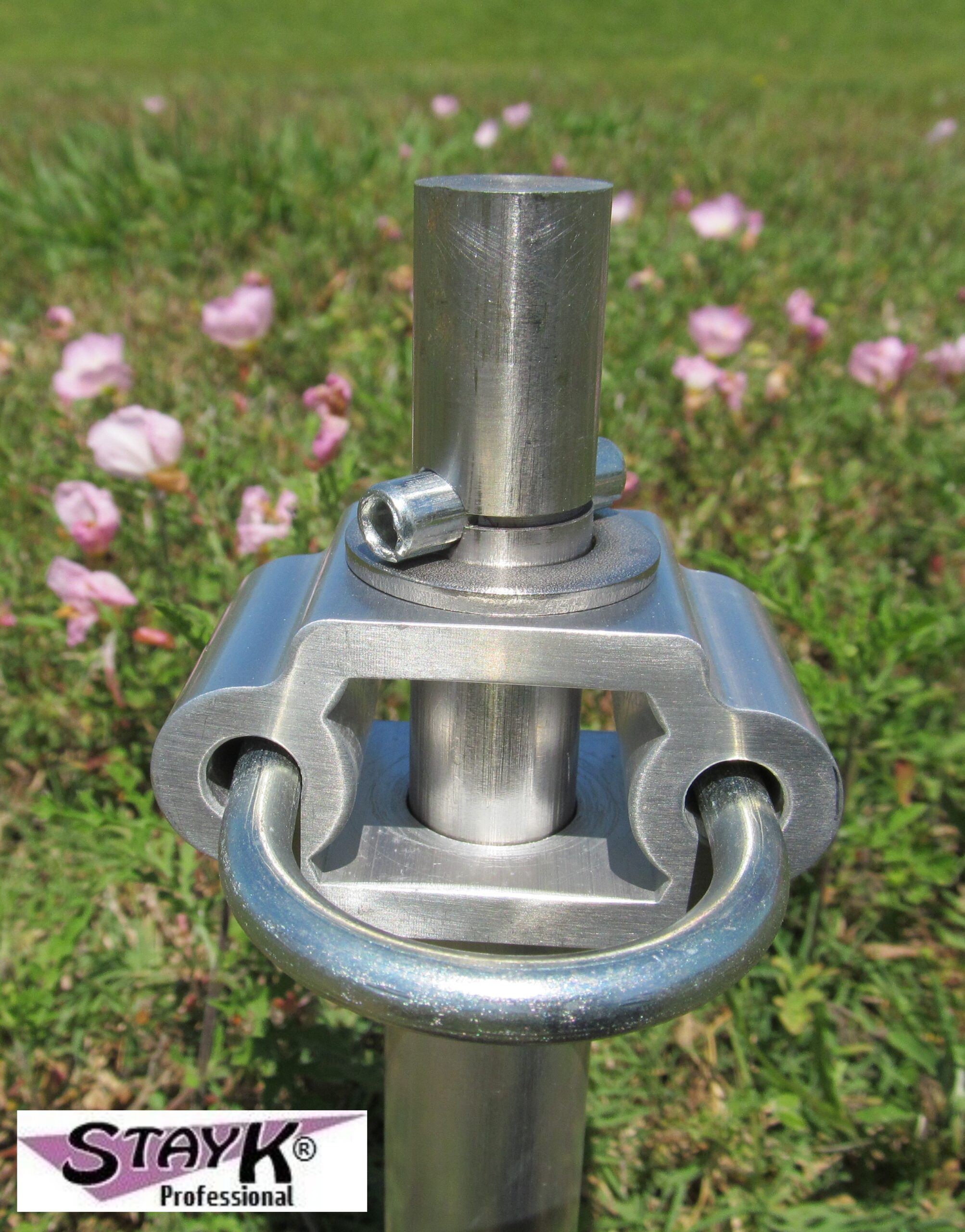 Close-up of a stainless steel connector with a ring and locking bolt, set against a background of green grass and pink flowers. Relevant for showcasing equipment durability.