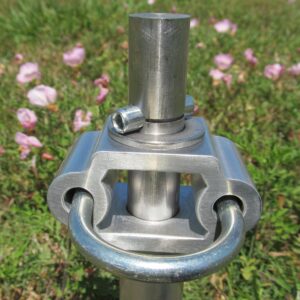 Metal connector with a loop and adjustable clamp, set against a background of pink flowers and green grass, likely for outdoor equipment usage.