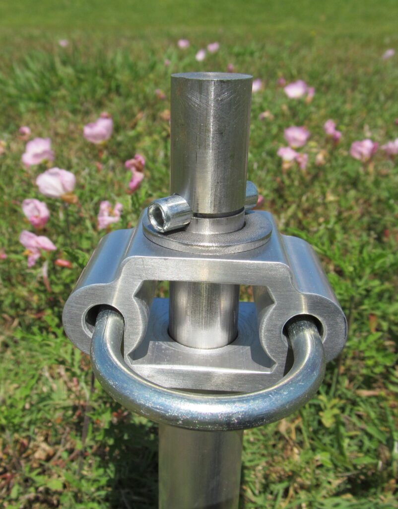 Metal connector with a loop and adjustable clamp, set against a background of pink flowers and green grass, likely for outdoor equipment usage.