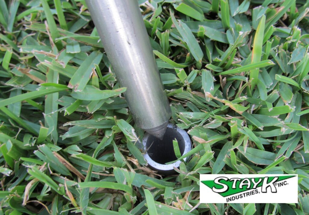 Metal rod positioned above a circular insert in grass, demonstrating the installation of a ground anchor by StayK Industries.