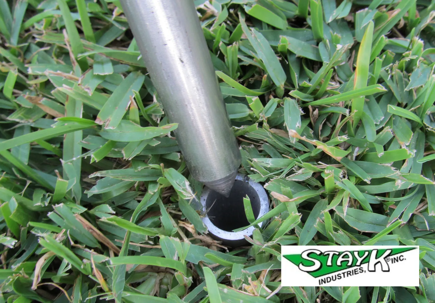 A metal tool positioned above a circular base set in green grass, likely used for ground installation. The tool's purpose relates to outdoor equipment.