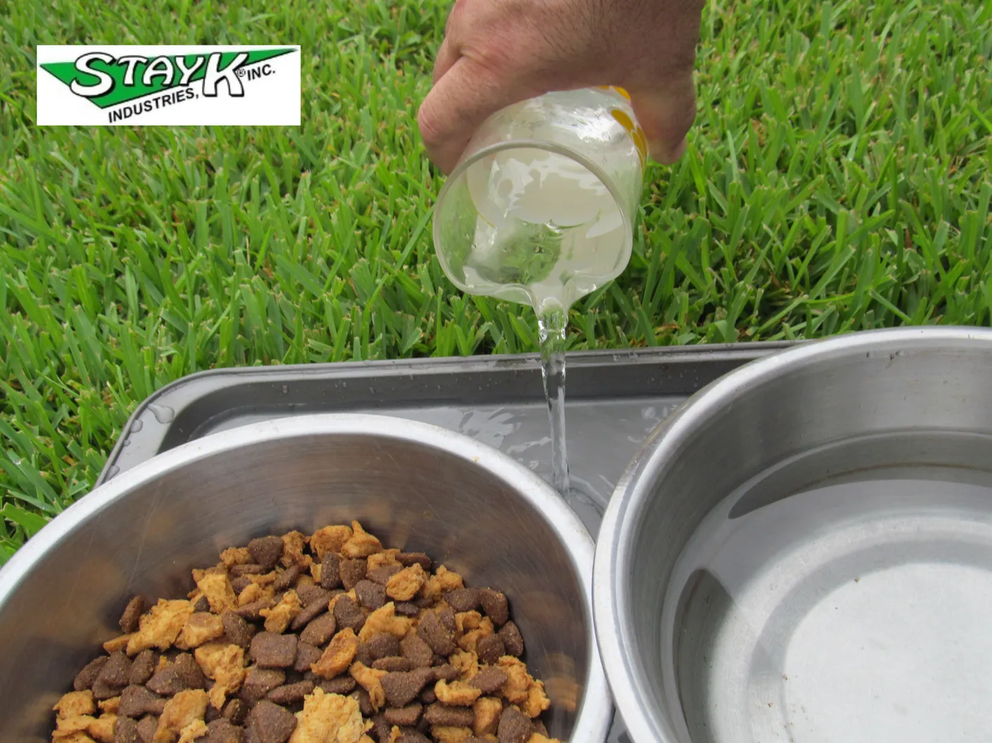 A hand pours water from a jar into a pet food bowl filled with kibble on green grass, highlighting pet care and hydration.
