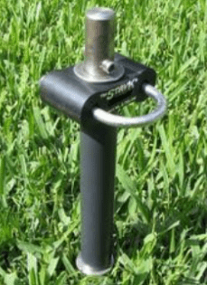 A metal ground anchor with a looped handle, designed for securing outdoor equipment or tents, positioned on green grass.