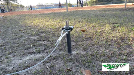 A ground anchor with a cable extends from it, set on a grass field near a baseball diamond, supporting sports equipment safety.