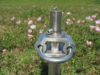 Metallic hitch pin is shown in focus against a backdrop of pink flowers in a grassy field, highlighting outdoor equipment and nature.